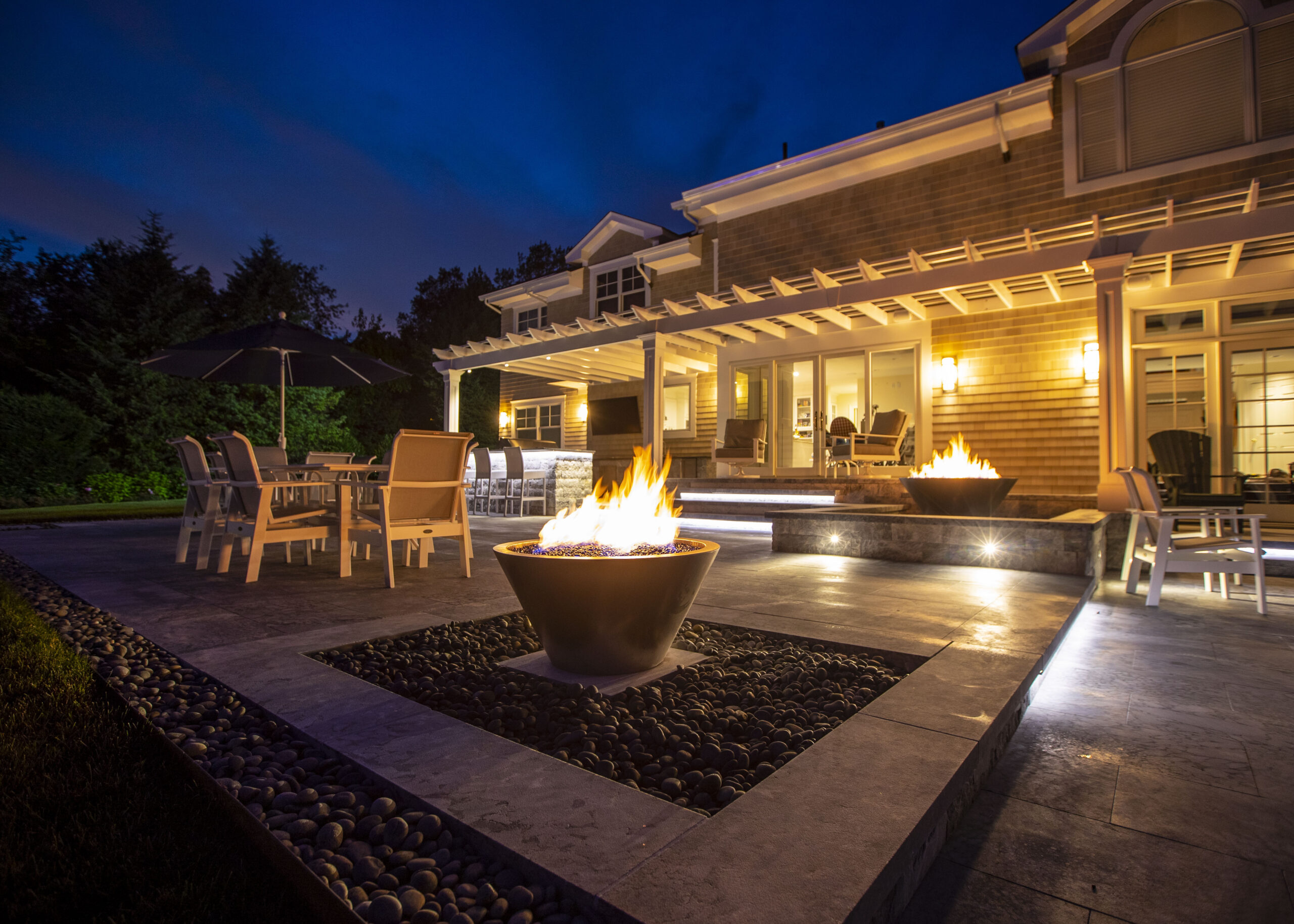 An illuminated patio and house exterior with dining furniture and fire bowls after dark.
