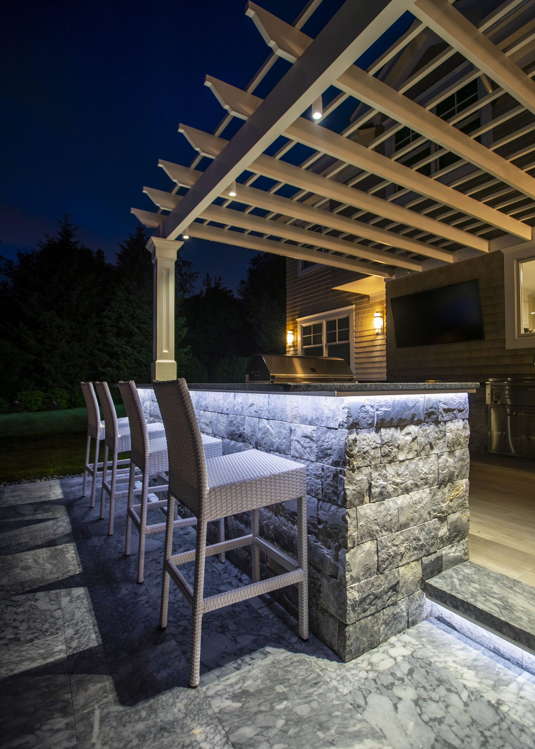 Lights beneath a white pergola above an illuminated stone bar beside a shingle-sided house.